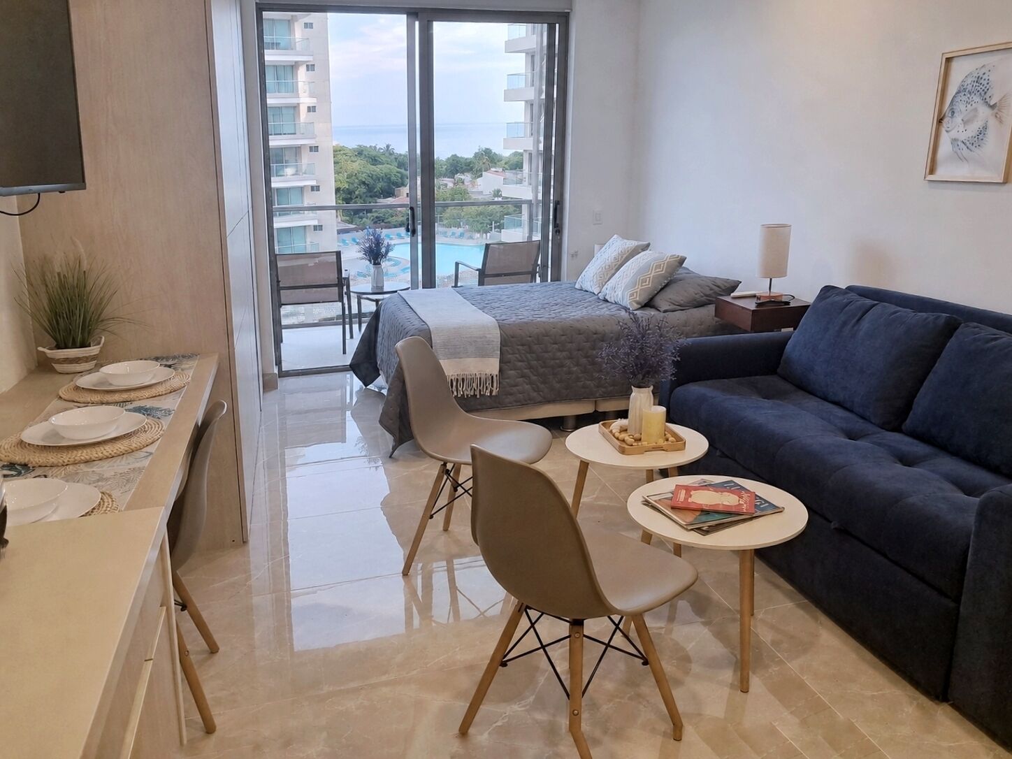 A modern hotel room with a balcony, a bed, a sofa, a dining area, and a TV.