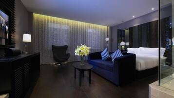 A modern hotel room with a bed, a sofa, a chair, a small table with a vase of flowers, and a desk with a lamp.