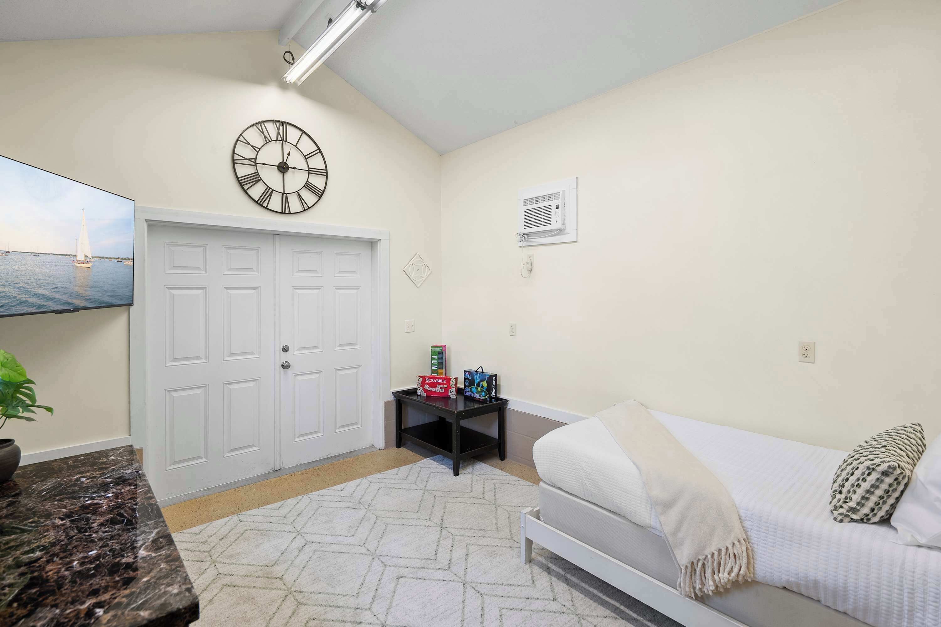 A bedroom with a bed, a wall clock, a TV, a small table with games, and a door.