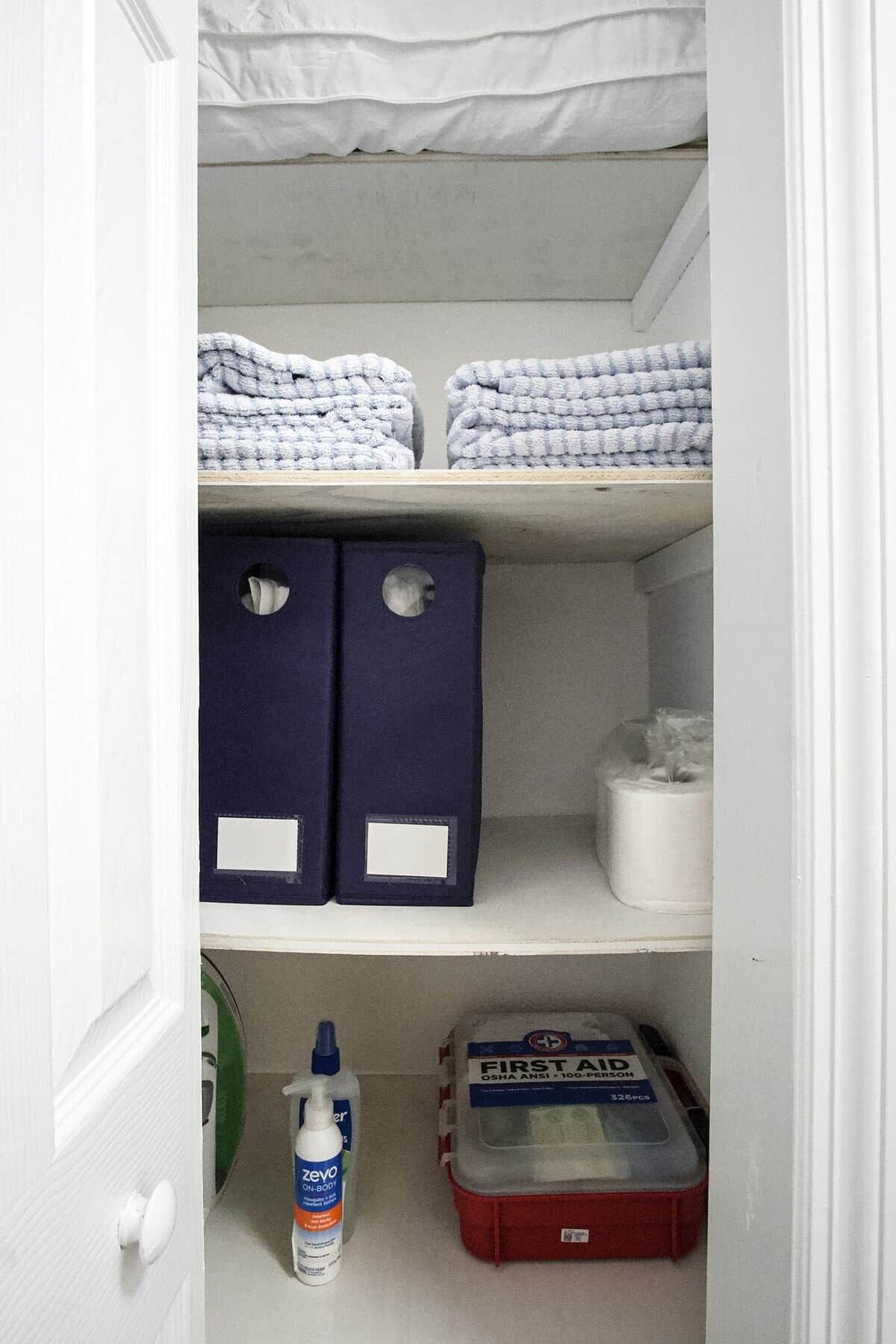 A closet with shelves containing folded towels, storage boxes, and a first aid kit.