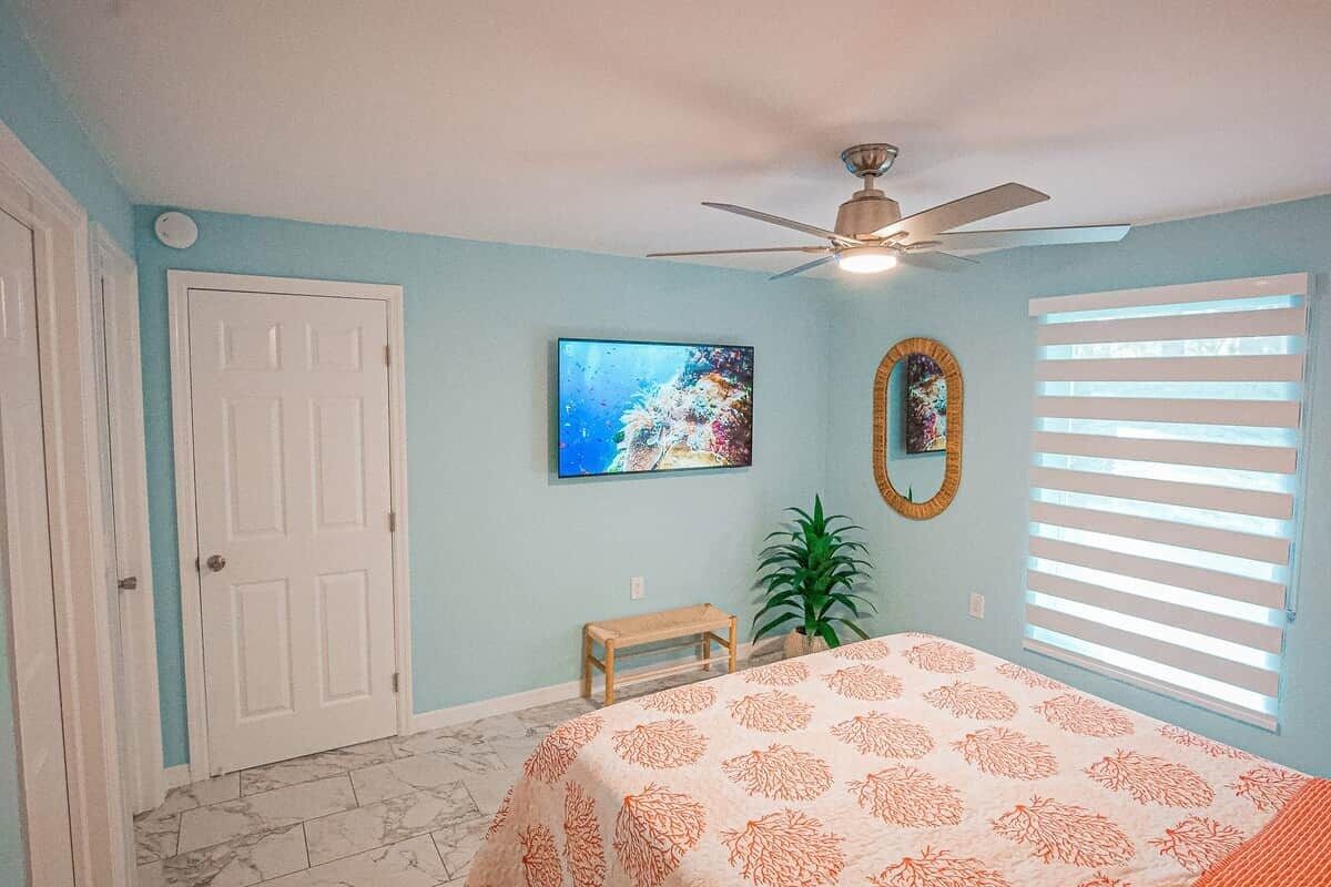 A bedroom with a bed, a ceiling fan, a window with blinds, a door, a painting, and a small bench.