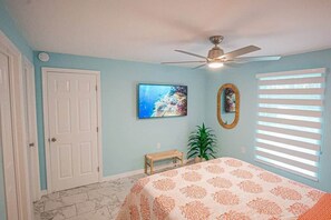A bedroom with a bed, a ceiling fan, a window with blinds, a door, a painting, and a small bench.