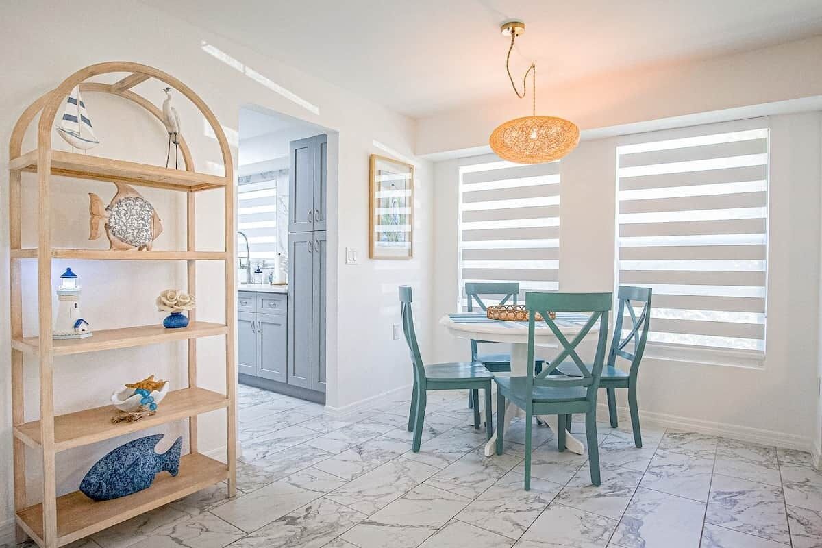 A dining area with a table and chairs, a wooden shelf with decorative items, and a window with blinds.