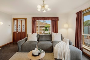 A cozy living room with a grey sofa, a small round table with candles, and a view of a garden through a window with brown curtains. - Modern 3BR Retreat | Near Launceston CBD (Youngtown)
