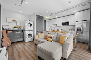 Ein modernes Apartment mit grauem Sofa, einer Waschmaschine, einer Gitarre und einer Küche mit weißen Schränken und Edelstahlgeräten.