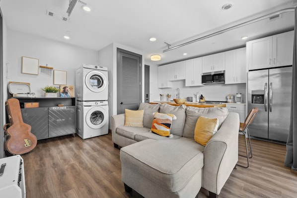 A modern apartment with a grey sofa, a washing machine, a guitar, and a kitchen with white cabinets and stainless steel appliances. - Downtown Condo · Rooftop + Gym · Walk to Broadway (Nashville)