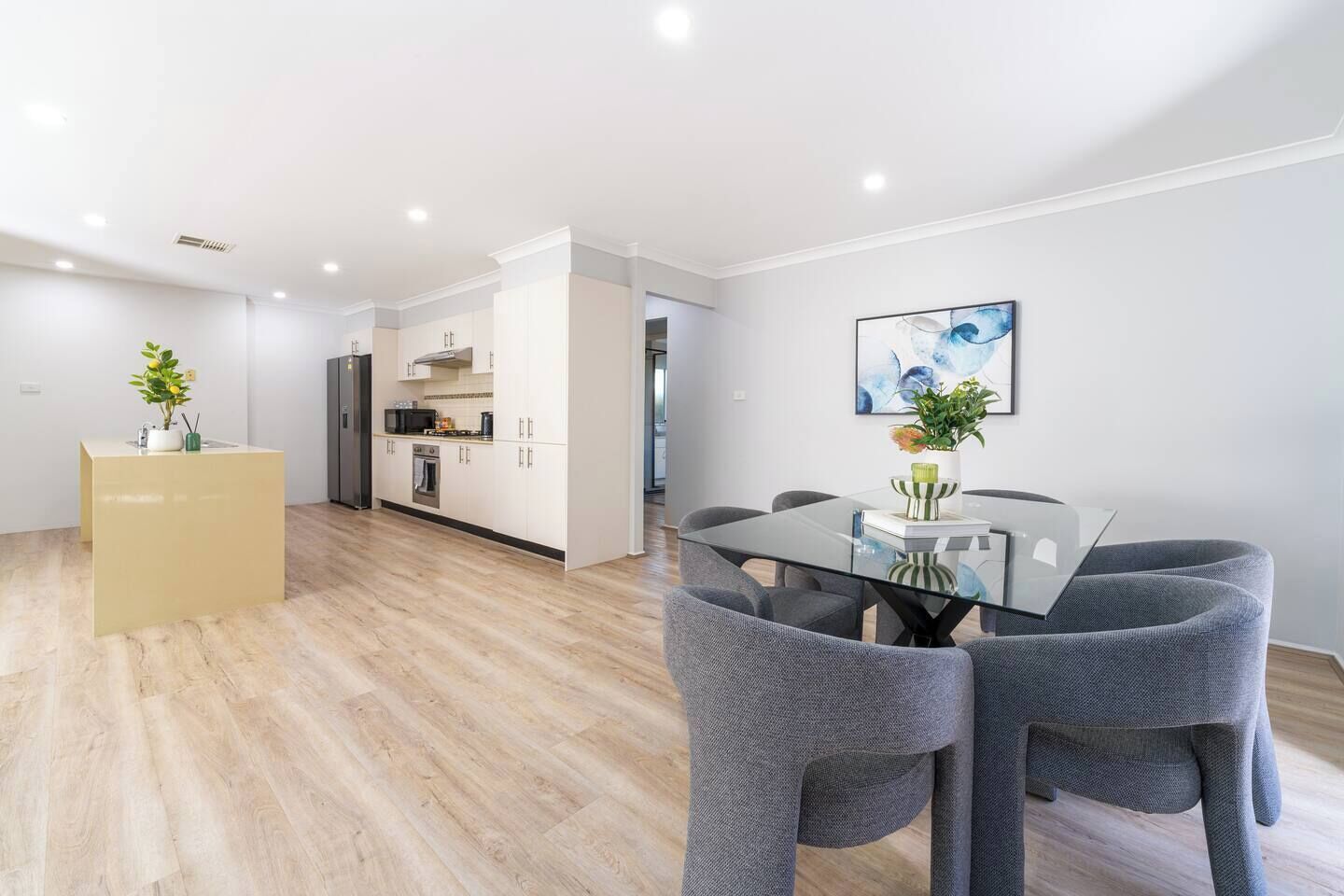 A modern kitchen with white cabinets, a central island, and a dining area with a glass table and grey chairs.