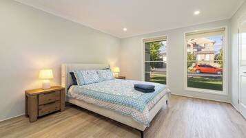 A bedroom with a bed, bedside lamps, a wooden nightstand, and a window with a view of a street and a car.