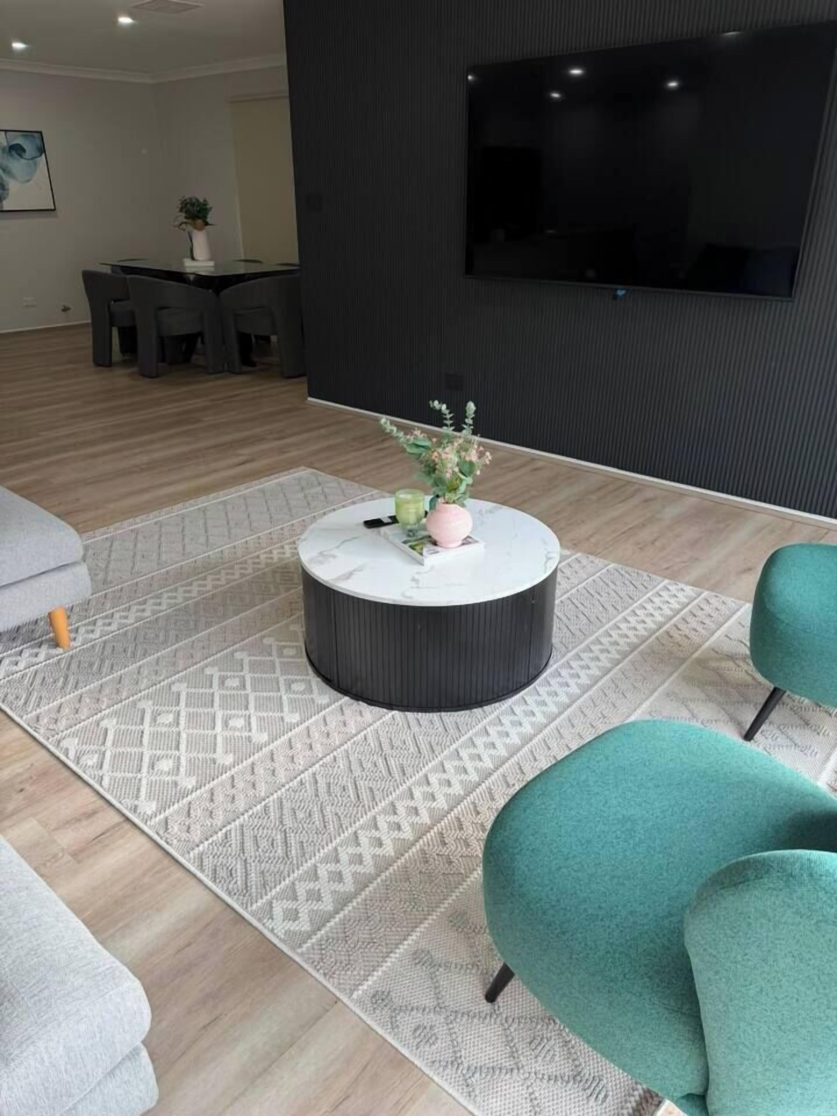 A modern living room with a round coffee table, teal armchairs, a TV mounted on the wall, and a dining area in the background.