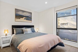 A bedroom with a bed, two bedside tables with lamps, a large window with blinds, and a painting on the wall. - Luxury on Gloucester (Christchurch)