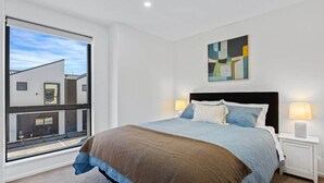 A modern bedroom with a large bed, two bedside lamps, a view of buildings through a window, and a piece of abstract art on the wall. - Luxury on Gloucester (Christchurch)
