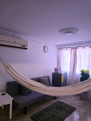 Ein Wohnzimmer mit einer Hängematte, einem Sofa, einem kleinen Tisch, einer Klimaanlage, einer Uhr, einem Fenster mit Vorhängen und einer Topfpflanze.