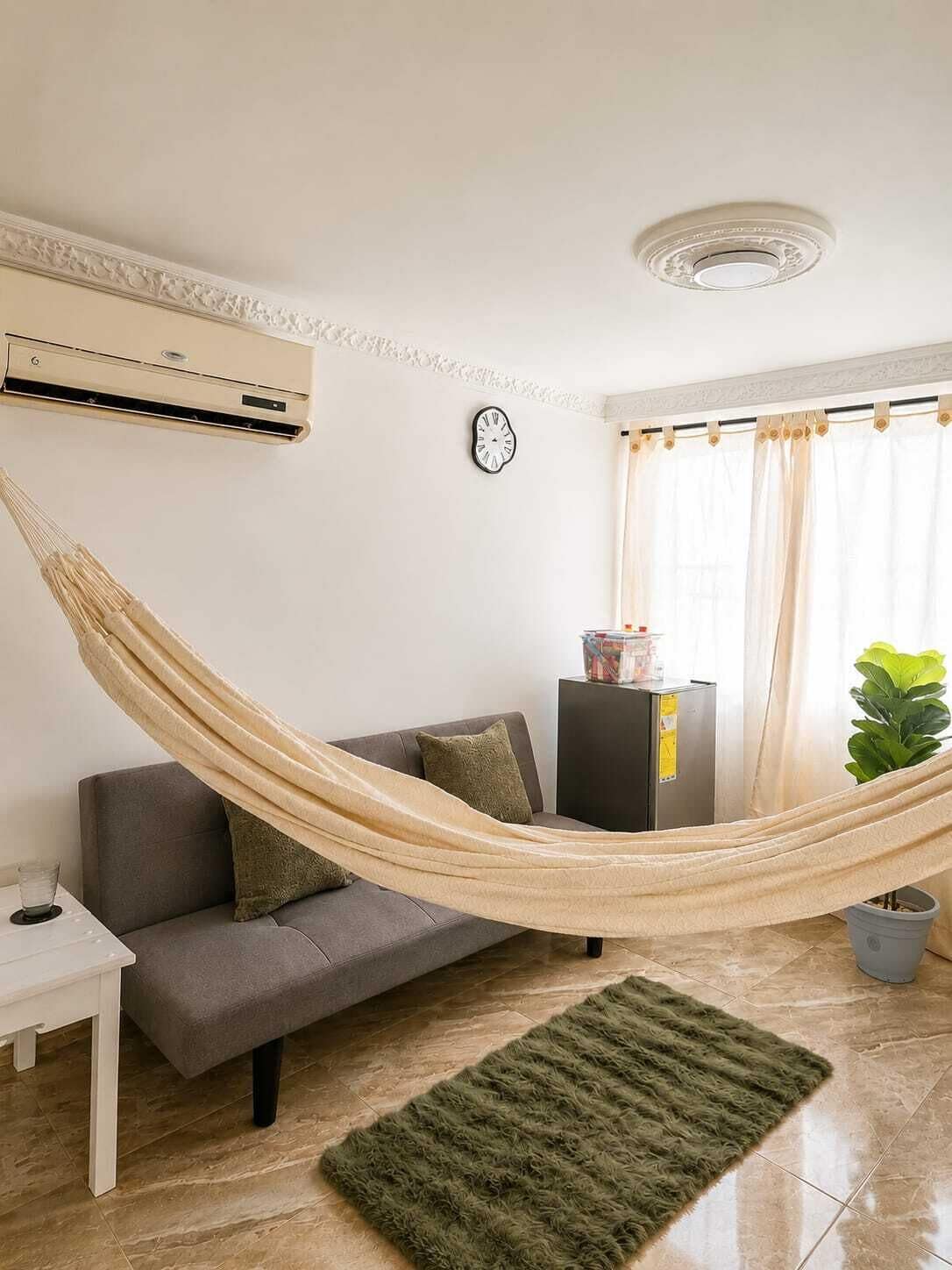 Ein Wohnzimmer mit einer Couch, einem kleinen Tisch, einem grünen Teppich, einer Hängematte, einer Uhr und einer Topfpflanze.