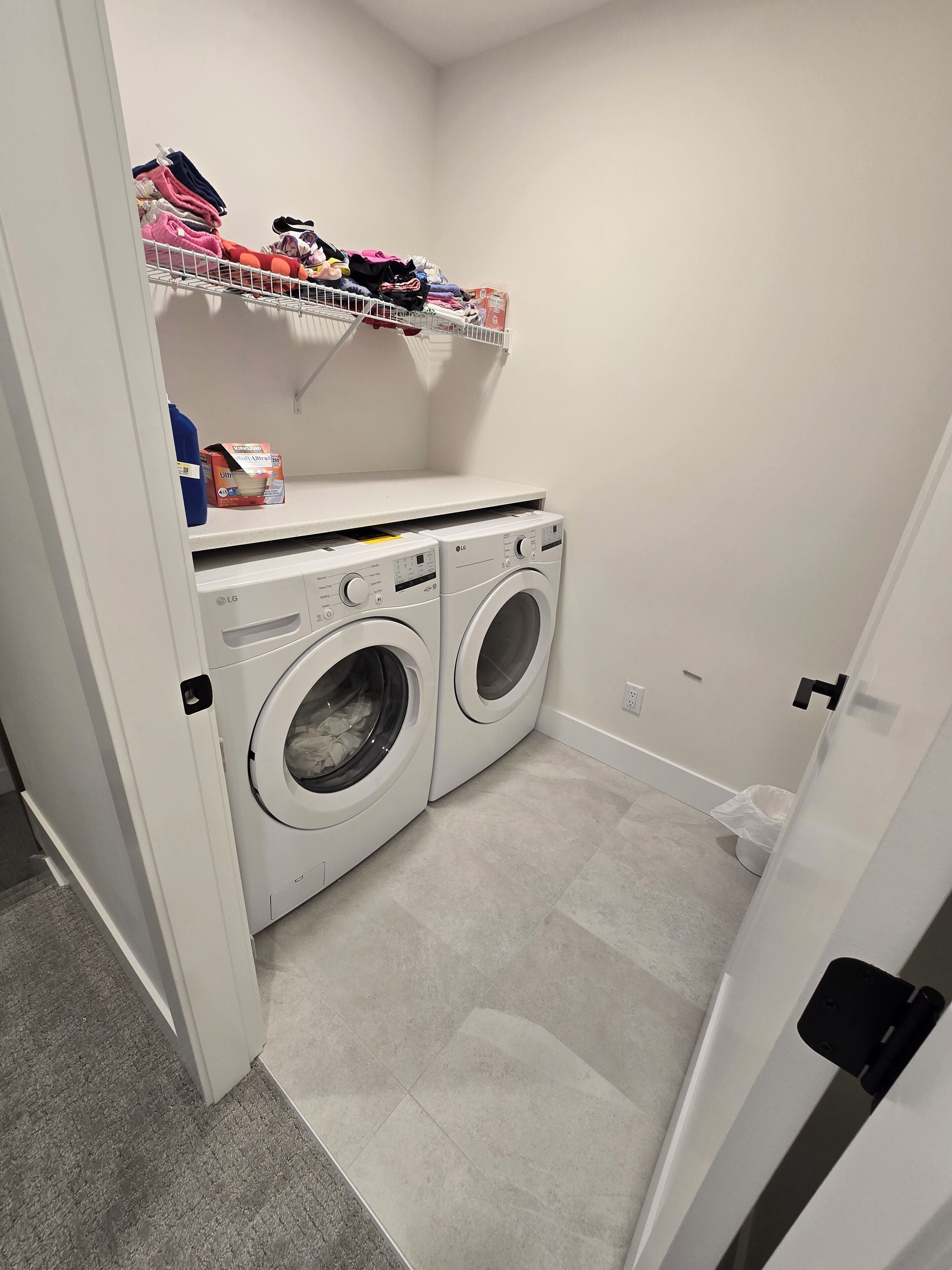 Une buanderie équipée de deux machines à laver LG à chargement frontal, blanches, d’une étagère où sont pliés les vêtements et d’une poubelle.