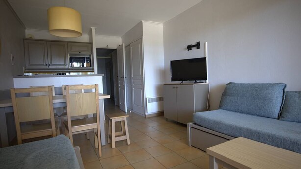 A compact living space with a kitchenette, a sofa, a TV, and a dining area with chairs. - 2-Bedroom Apartment in Les Rivages des Issambres (LES ISSAMBRES)