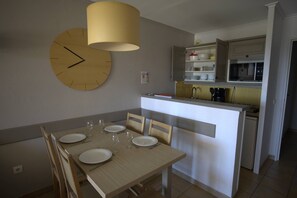 A dining area with a wooden table set for four, a large wall clock, and a kitchen in the background. - 2-Bedroom Apartment in Les Rivages des Issambres (LES ISSAMBRES)