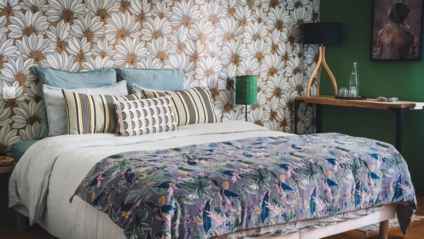 Quarto com colcha de padrão floral, mesa lateral verde com abajur e parede com papel de parede com padrão de folhas.