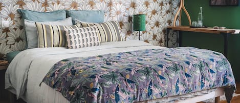 Une chambre à coucher avec une couvre-lit à motifs floraux, une table de chevet verte avec une lampe, et un mur recouvert d’un papier peint à motifs de feuilles.