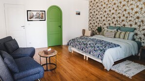 Um quarto com uma cama, uma porta verde, uma mesa redonda pequena com uma caixa de lenços, um sofá cinzento e um papel de parede estampado.