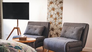 Quarto com duas poltronas cinzentas, uma pequena mesa de apoio de madeira, uma TV preta em suporte de tripé e um papel de parede estampado.