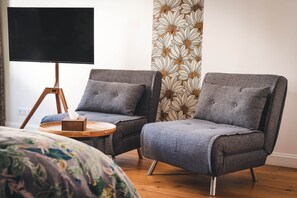 A room with two grey armchairs, a small wooden side table, a black TV on a tripod stand, and a patterned wallpaper. - Les Sentiers de Solutré Room (FUISSE)