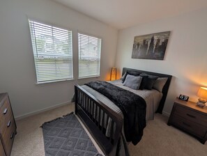 Een slaapkamer met een bed, een nachtkastje, een lamp, een schilderij aan de muur en twee ramen met jaloezieën.