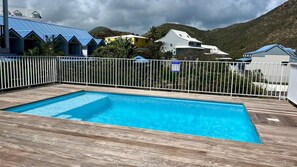 A rectangular swimming pool with a wooden deck, surrounded by a white fence, set against a backdrop of houses and a mountainous landscape. - The Cherry   unit 1BR 1.5BTH near Pinel Island (SAINT MARTIN)