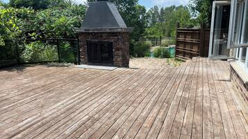 A wooden deck with a stone fireplace, surrounded by greenery and a swimming pool.