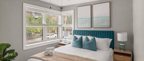 A bedroom with a bed, a ceiling fan, a window with blinds, a bedside table with a lamp, and artwork on the wall.