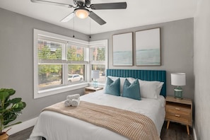 A bedroom with a bed, a ceiling fan, a window with blinds, a bedside table with a lamp, and artwork on the wall.