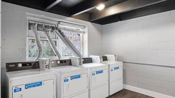 A laundry room with four white washing machines, a vent system, and a window with a view of buildings.