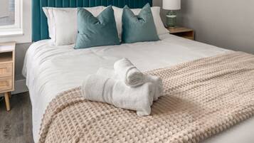 A neatly made bed with a white duvet, teal and white pillows, a beige textured throw blanket, and a white towel rolled up on the bed.