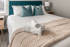 Una cama bien tendida con un edredón blanco, almohadas turquesa y blancas, una manta beige con textura y una toalla blanca enrollada sobre la cama.