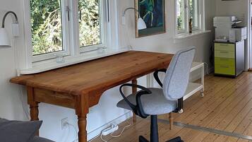 Een kamer met een houten bureau, een grijze bureaustoel, een raam met uitzicht op groen, een printer en een klein kastje.