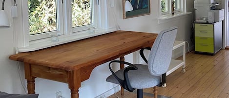 Habitació amb escriptori de fusta, cadira de oficina gris, finestra amb vista a vegetació, una impressora i un petit armari.
