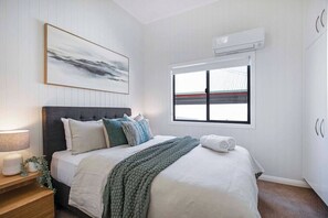Ein Schlafzimmer mit Bett, Nachttisch, Lampe, Klimaanlage, Fenster und einem Kunstwerk an der Wand.