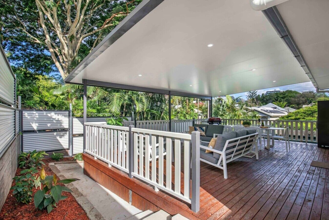 A covered outdoor deck with a white railing, a seating area with cushions, and a view of the surrounding greenery.