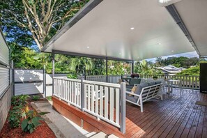 A covered outdoor deck with a white railing, a seating area with cushions, and a view of the surrounding greenery. - Central Retreat - 4Bed (Petrie Terrace)