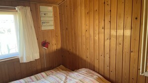 A room with wooden walls, a bed with a striped bedspread, a window with a white curtain, and a red lamp on the wall. - Beautifully situated classic wooden summercottage (Hadsund)