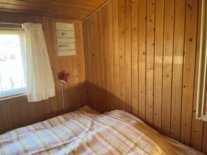 A room with wooden walls, a bed with a striped bedspread, a window with a white curtain, and a red lamp on the wall. - Beautifully situated classic wooden summercottage (Hadsund)