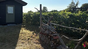 A rustic wooden cabin with a door and a window, surrounded by a garden with a pile of firewood and a wooden fence. - Beautifully situated classic wooden summercottage (Hadsund)