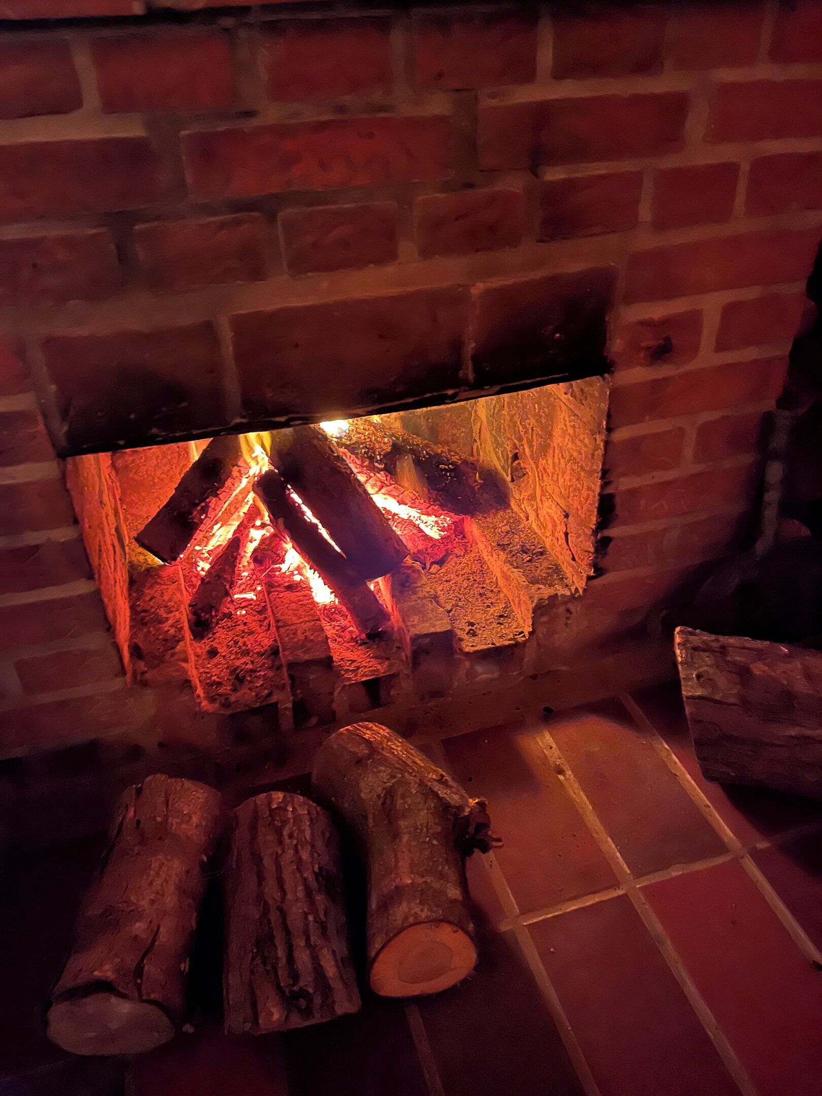 A fireplace with logs burning, a brick wall, and a tiled floor.