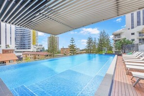 Ein Schwimmbecken mit einer Holzterrasse und Liegestühlen, umgeben von Gebäuden und Bäumen.