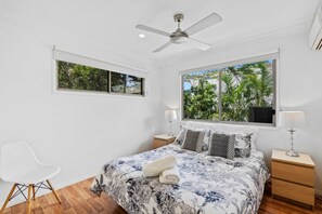 A bedroom with a bed, a ceiling fan, a window with a view of greenery, and a white chair.