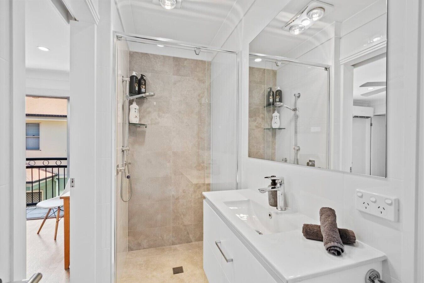 Une salle de bain moderne avec une douche à l’italienne en verre, un meuble-lavabo blanc doté d’un grand miroir et une serviette posée sur le plan de travail.