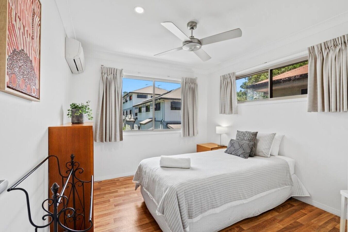 A bedroom with a bed, a ceiling fan, a window with curtains, a bedside table, and a view of buildings outside.