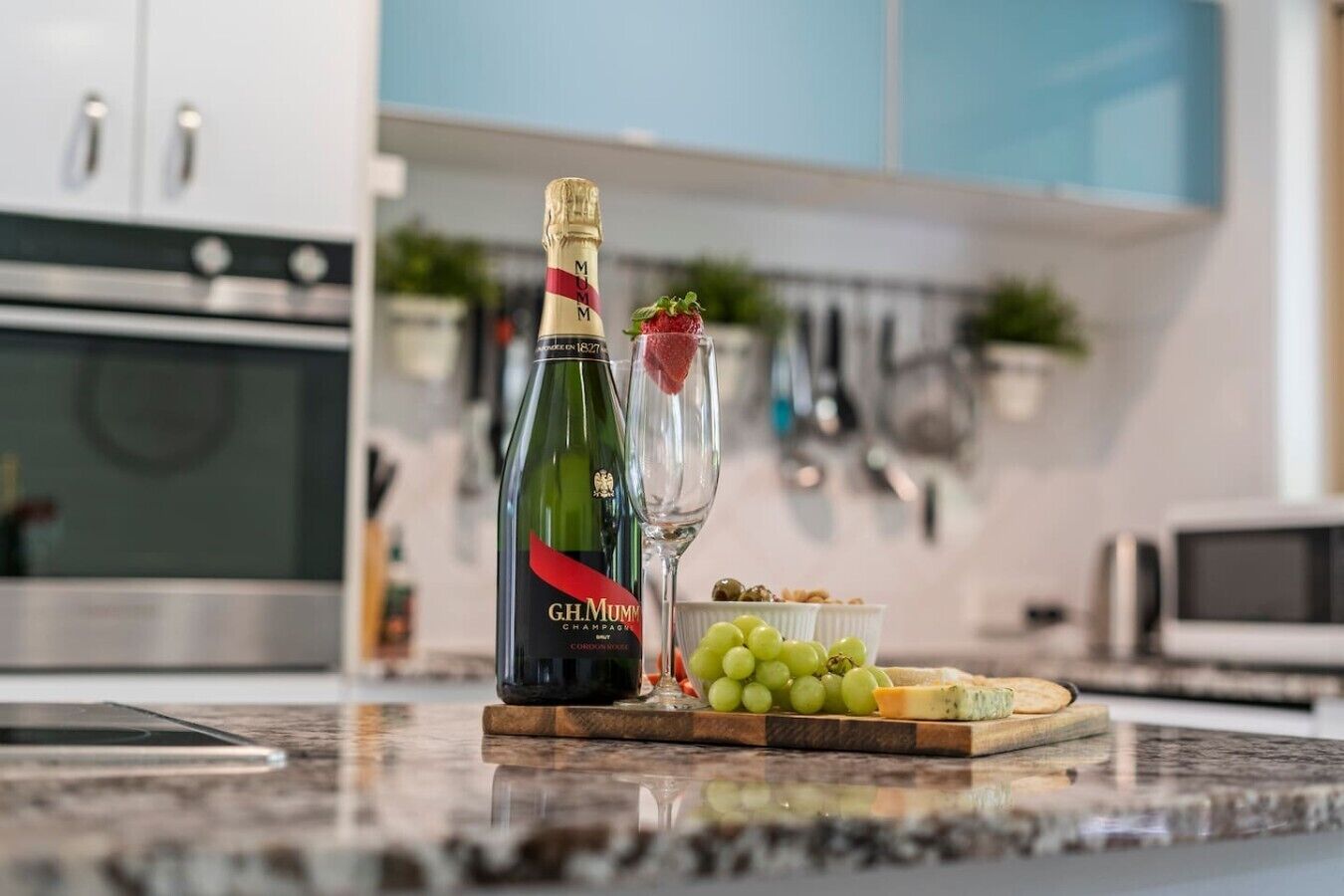 A bottle of G.H. Mumm champagne, a glass with a strawberry, grapes, and cheese on a wooden board in a modern kitchen.