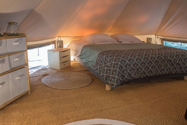 Une chambre d’hébergement sous tente comprenant un lit, une table de chevet en bois avec des tiroirs, une lampe et un meuble de rangement doté de plusieurs compartiments.
