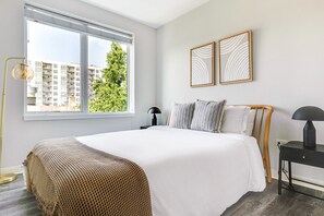 A modern bedroom with a large bed, a bedside table, a floor lamp, and a window with a view of buildings and trees. - Blueground | Edgewater, gym, nr Lake Michigan (Chicago)