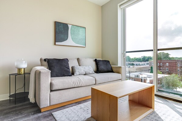 Een moderne woonkamer met een bank, een salontafel, een bijzettafeltje met een lamp, een ingelijst abstract schilderij en een groot raam met uitzicht op de stad.